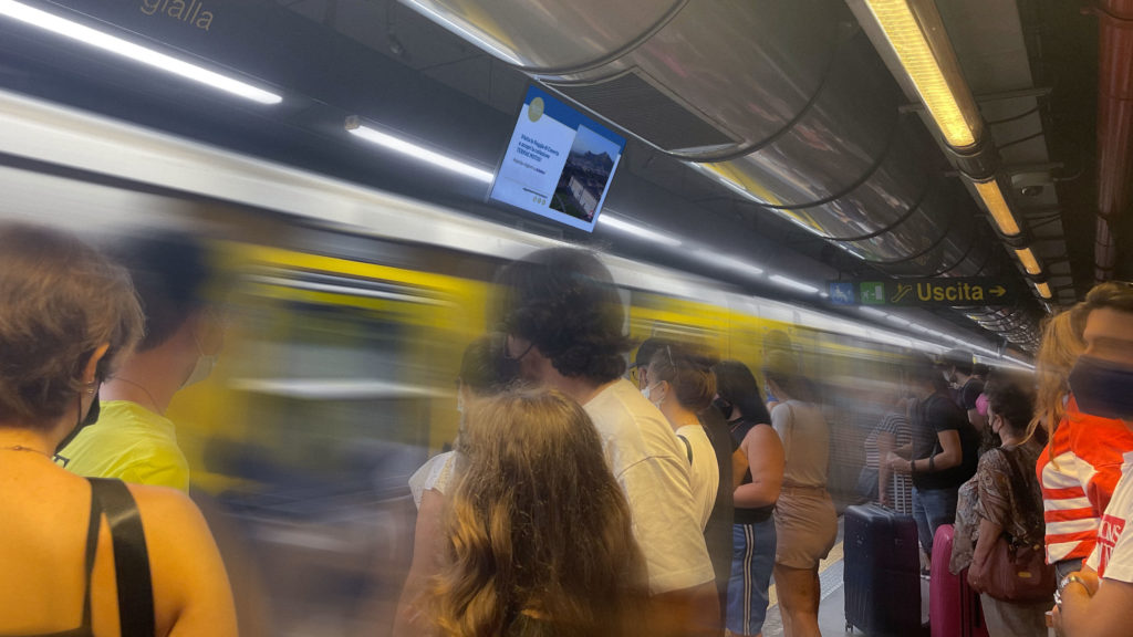 Viaggiatori sulla banchina della metropolitana di Napoli con monitor VNN.it